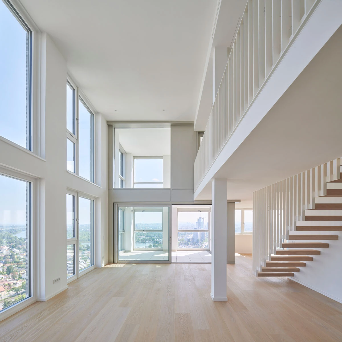 Empty two-story contemporary living room with white walls, light wood floor, and an open white staircase; large windows frame a city view.