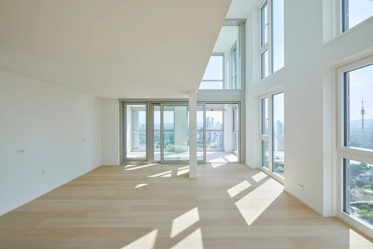 Bright empty living room with white walls, light wood floor, and floor-to-ceiling windows revealing a city skyline.