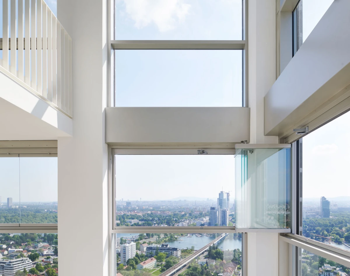 Modern high-rise interior with large floor-to-ceiling windows; an open glass door overlooks a city skyline and river.