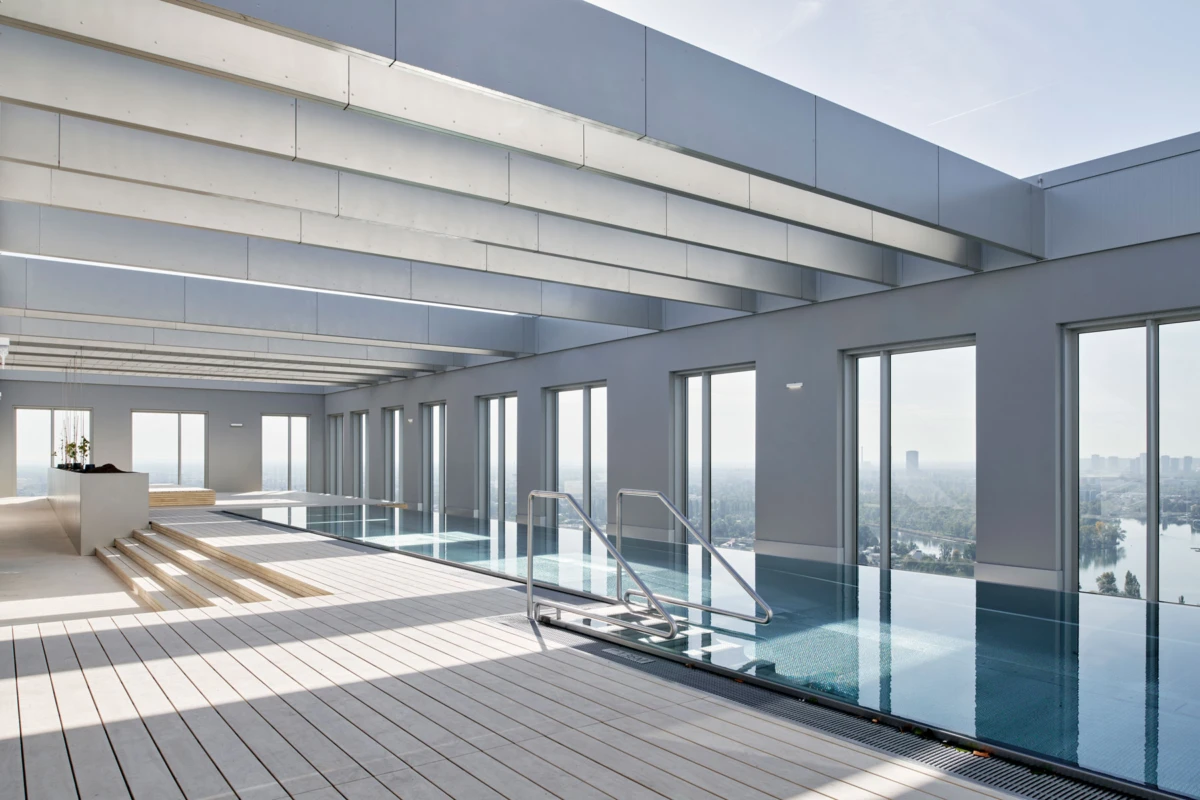 Indoor rooftop pool, unoccupied, with wooden deck, stainless steel handrail, and floor-to-ceiling windows; city skyline visible.