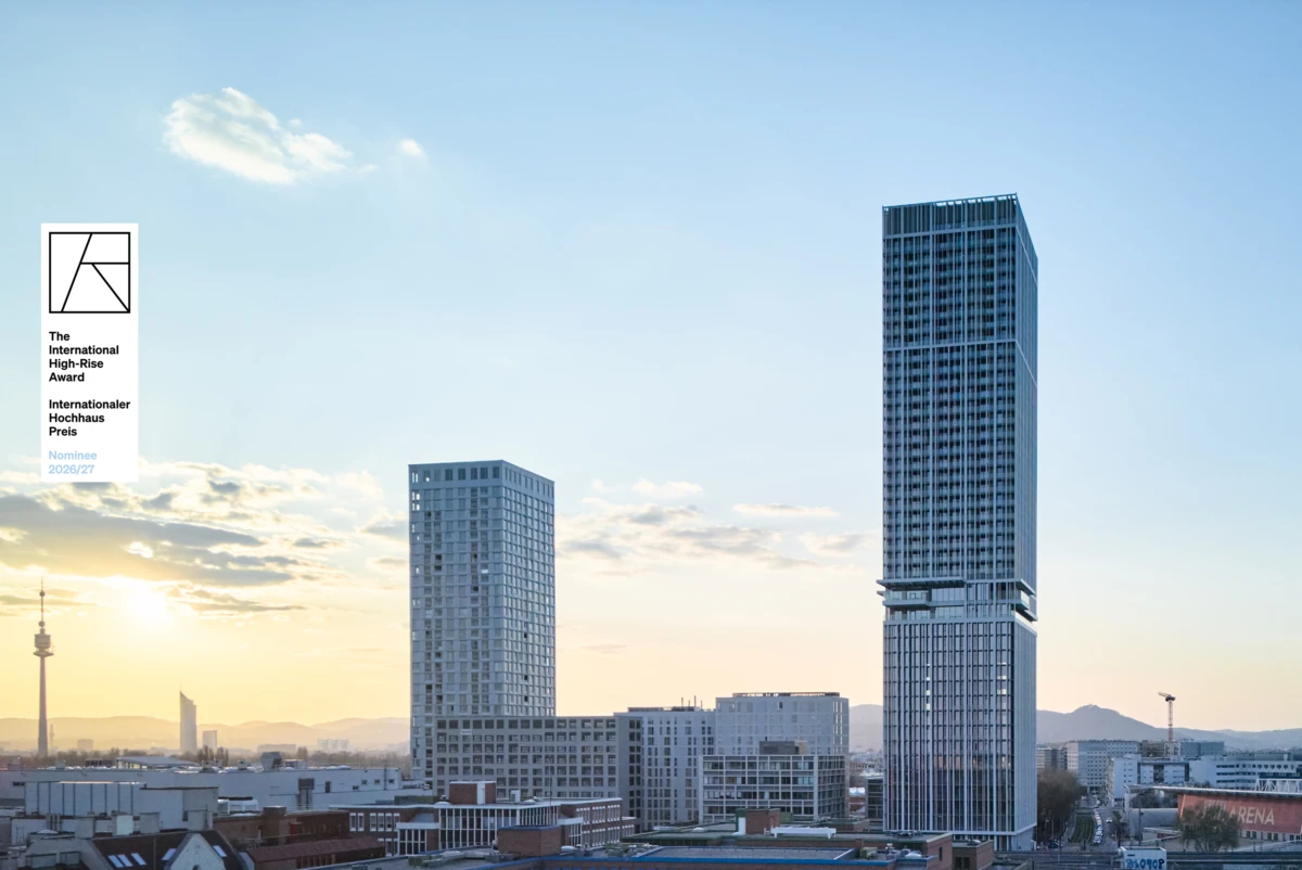 Sunset skyline with a tall glass-clad skyscraper on the right and a shorter rectangular high-rise to its left; a plaque on the left foreground.