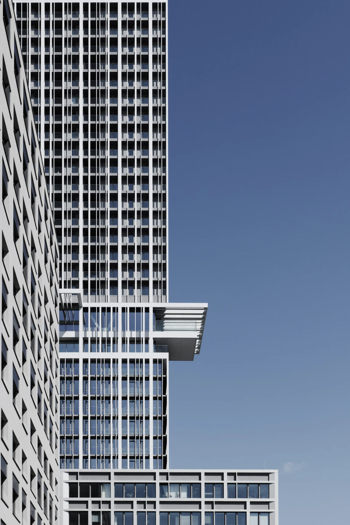 Tall modern glass office tower with a grid of windows and a cantilevered platform extending from its midsection against a clear blue sky.