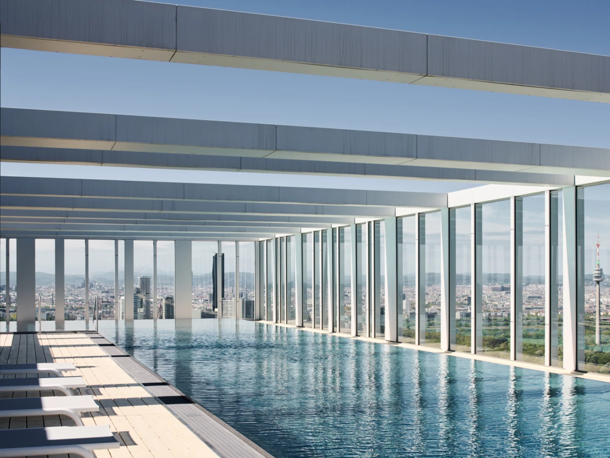 Rooftop infinity pool with glass walls overlooking a city skyline; white lounge chairs line the pool deck.