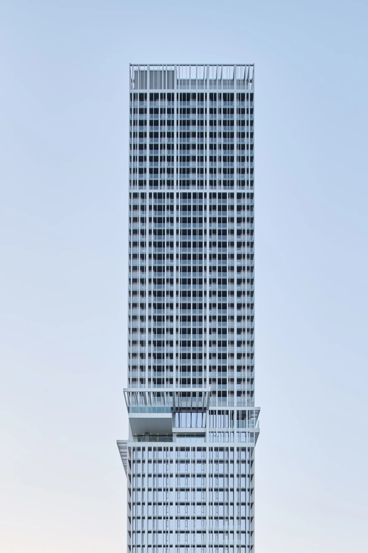 Tall modern skyscraper with stepped setbacks and a grid-like facade against a pale blue sky.