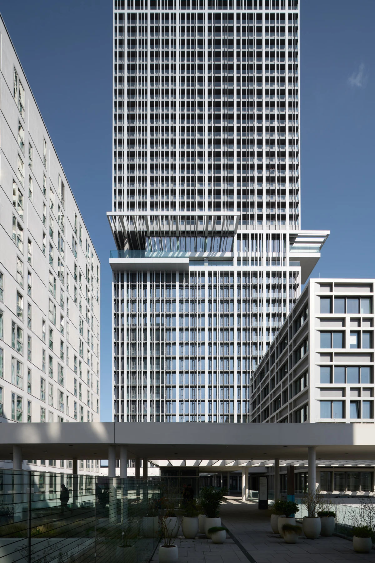Towering glass-clad skyscraper with stacked terraces, framed by adjacent modern buildings; pedestrian plaza with planters and a covered walkway.