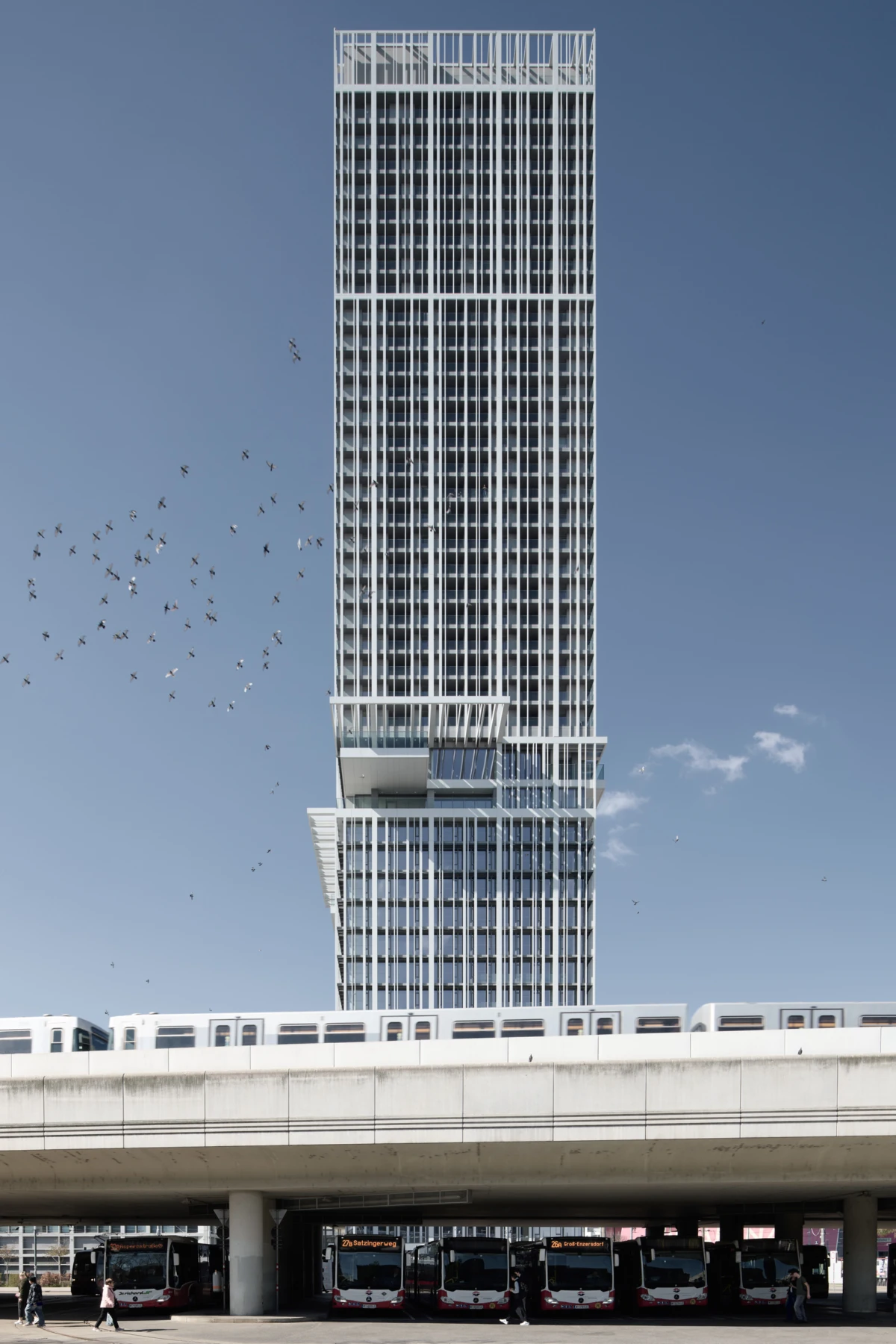 Glass-clad skyscraper with a stepped lower section; flock of birds in the sky; elevated transit deck with buses beneath in a city setting.