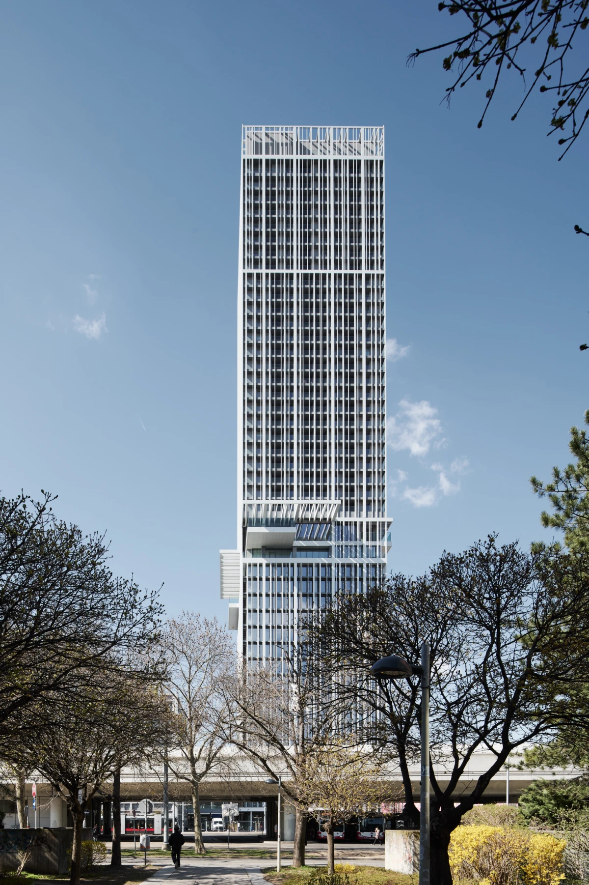Tall glass high-rise with a grid facade rises above a park-lined street; leafless trees and pedestrians in the foreground.