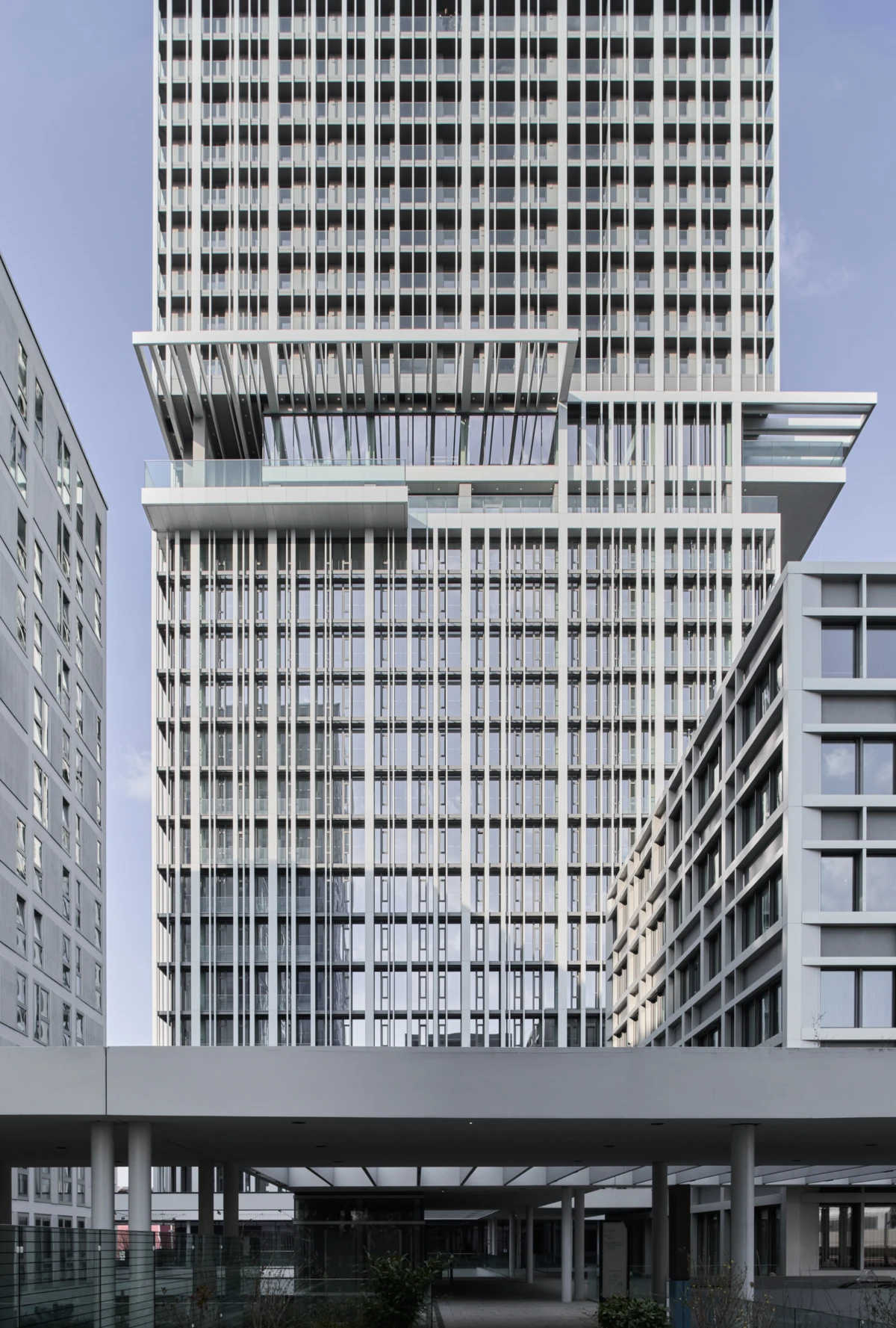 Tall glass skyscraper rising with a grid of windows and stacked terraces, in an urban complex with a covered pedestrian walkway.
