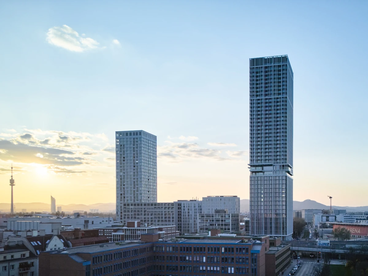 Two glass high-rise towers dominate a modern urban skyline at sunset, with low-rise buildings and distant hills.