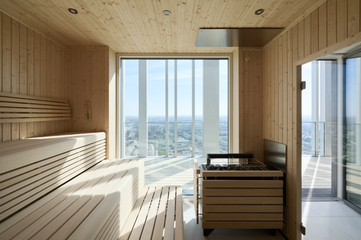 Interior of a wooden sauna with two-tier benches and a metal heater, large floor-to-ceiling window showing a cityscape.