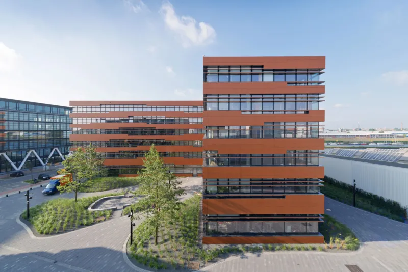 Multi-story red-brick office building with wide glass windows, set in a landscaped courtyard with paved walkways.