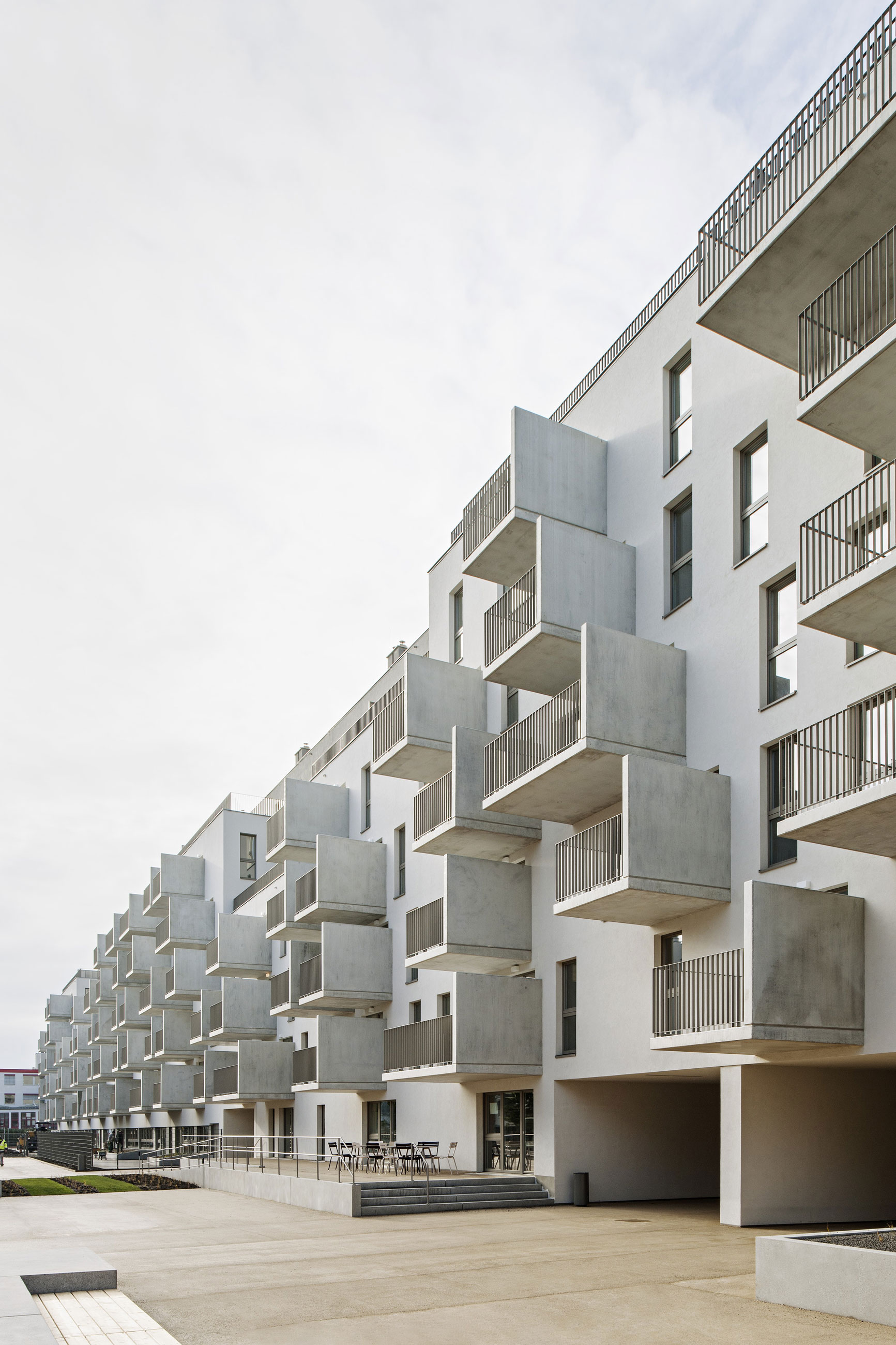 Modern white apartment building with stacked concrete balconies along an open plaza; ground-floor seating outside.