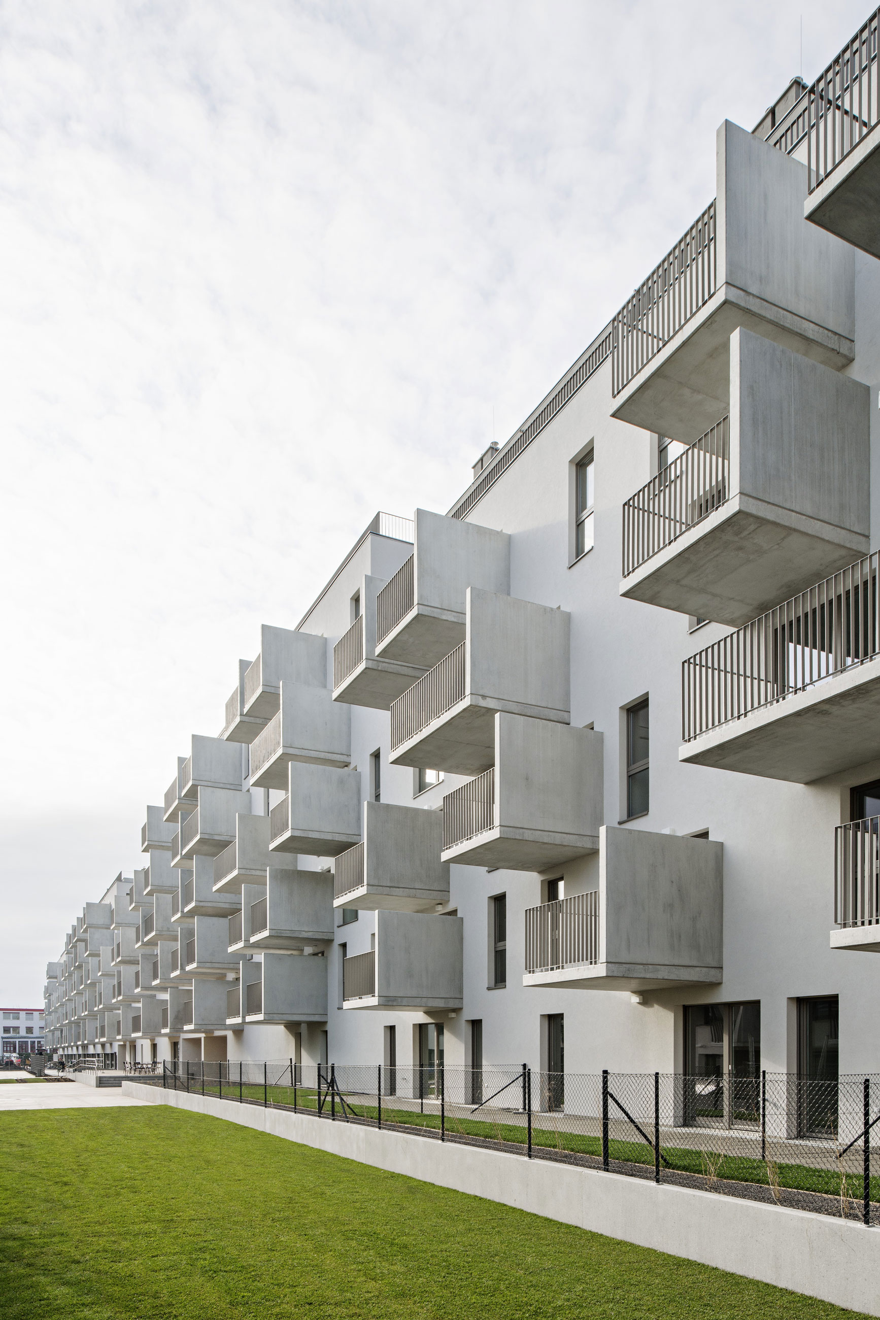 Modern white apartment building with protruding concrete balconies along a grassy lawn and chain-link fence, viewed from the side.