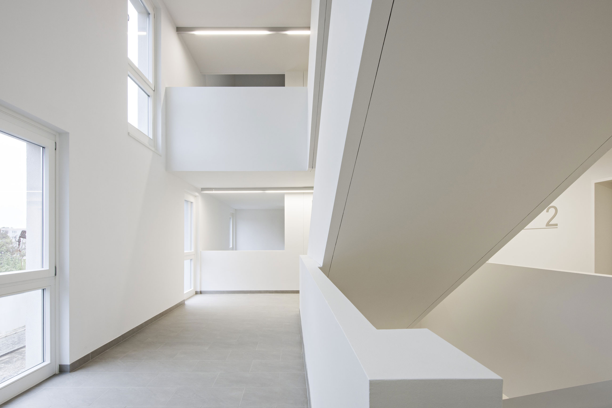 Bright white modern building interior with an open atrium, upper-level balconies, and a large diagonal staircase.