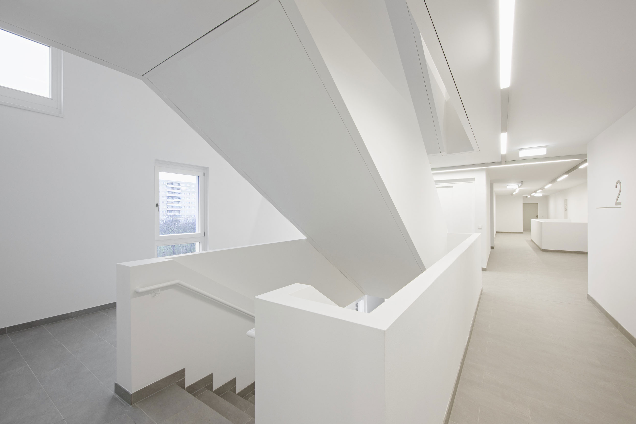 Angular white staircase dominates a bright, minimalistic interior hallway in a modern building; gray tiled floor, white walls, and distant corridor.