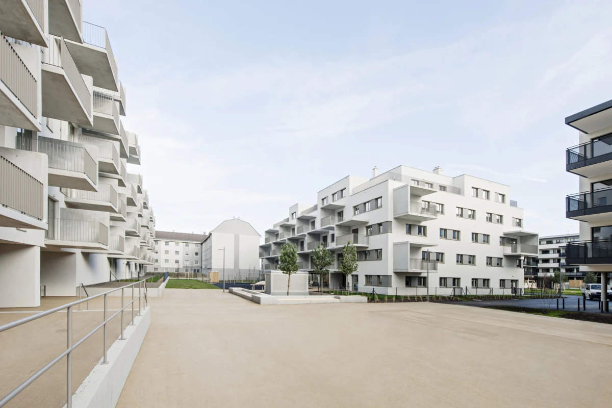 Modern white apartment buildings enclose a spacious courtyard with a paved plaza, balconies, and small trees.