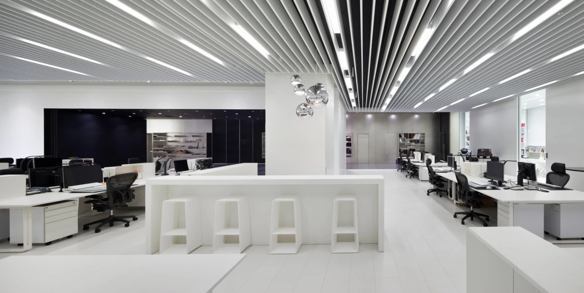Open-plan office with white desks and black chairs, a central white counter with four stools, and a striped ceiling with pendant lights.