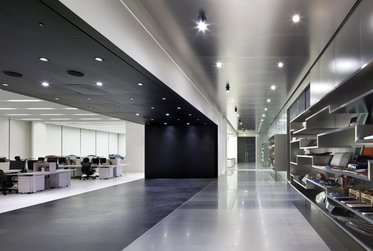 Modern office interior: open-plan workspace on the left, glossy corridor with shelving on the right, black wall at center.