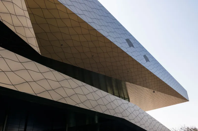 Cantilevered modern building with a diamond-patterned metallic facade extending over a dark base, set against a pale sky.