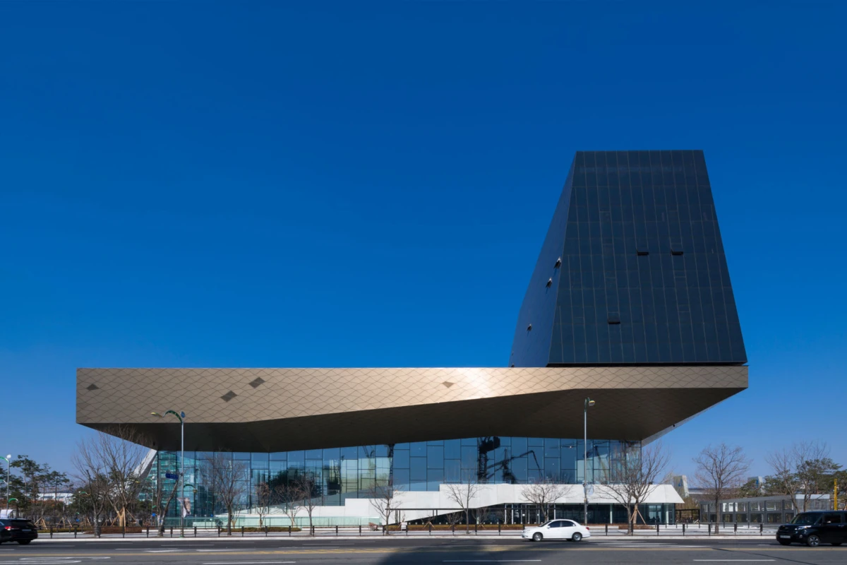 Contemporary building with a cantilevered metal overhang and a tall dark glass tower, along a glass-front facade; street with leafless trees and cars.