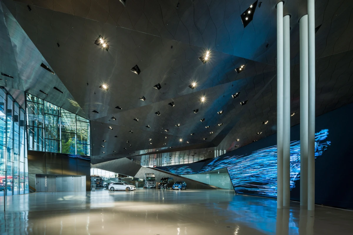 Futuristic car showroom interior with glass walls, glossy floor, angular dark ceiling, and several cars on display beside a blue wave light wall.