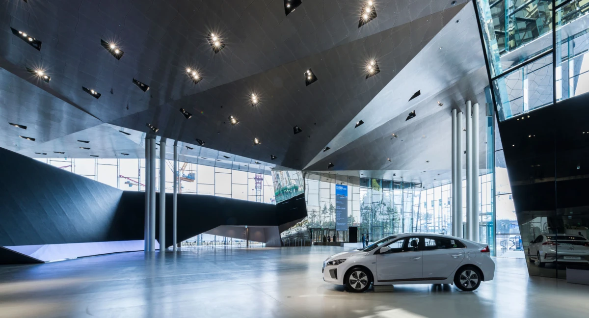 White sedan displayed on a polished showroom floor inside a futuristic automotive showroom with glass walls and an angular metallic ceiling.