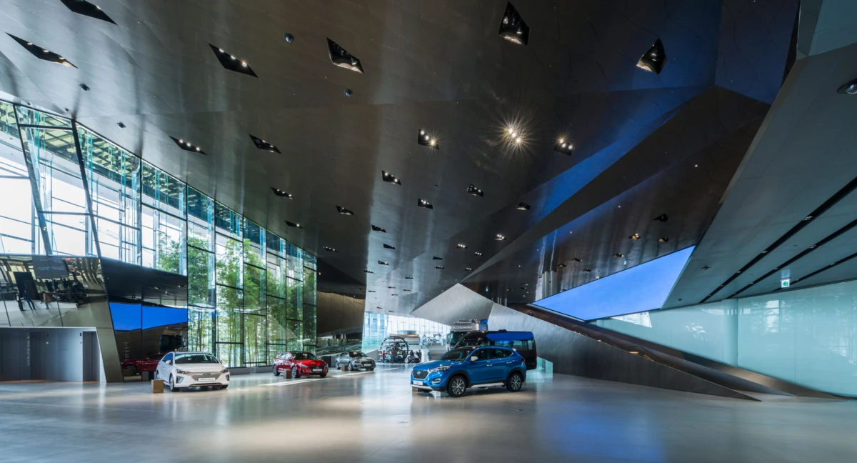 Cars on display inside a modern car showroom with glass walls, a polished floor, and a dark angular ceiling.