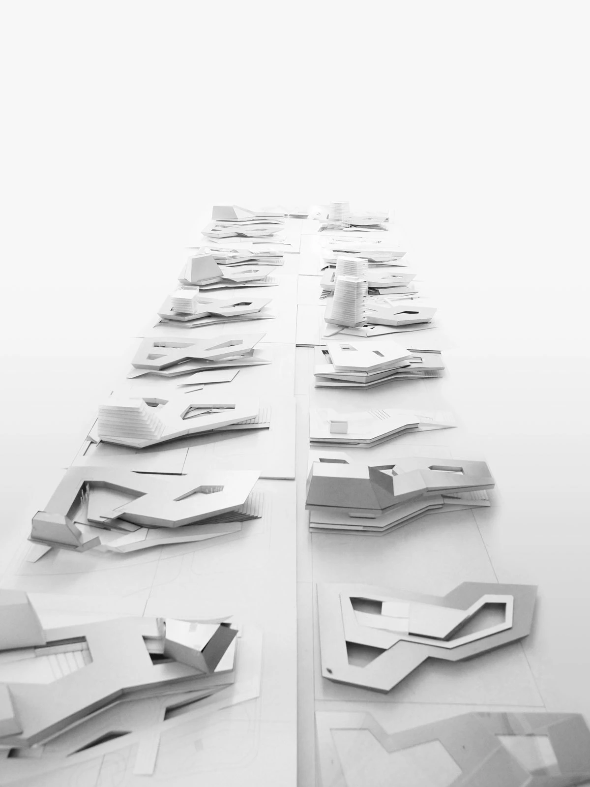 Two long rows of white architectural models arranged on a bright white studio table, viewed from end to end.