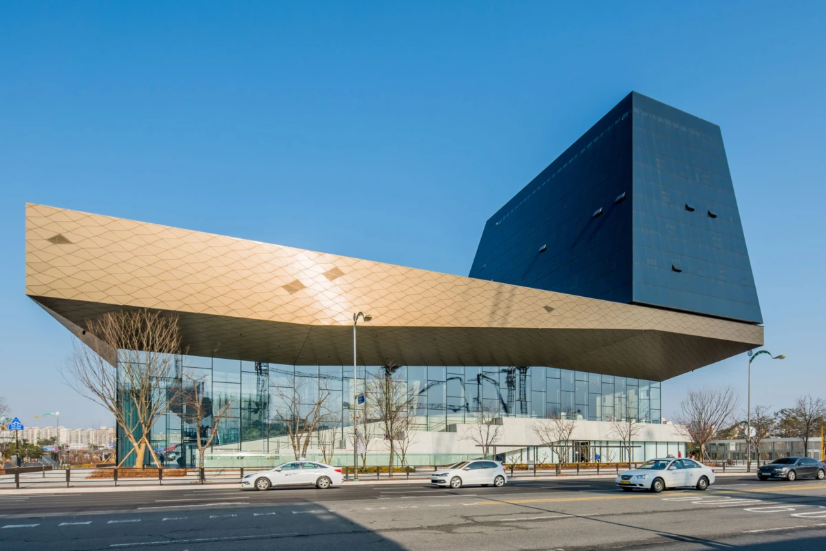 Modern cultural center with glass facade and a beige cantilever, topped by a blue rectangular tower, along a busy city street with cars.