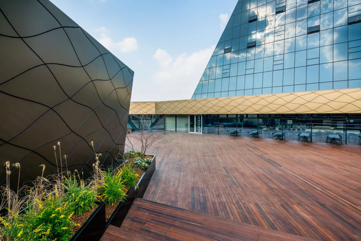 Wooden terrace between angular black wall with curved lines and a glass-clad pyramid building; planters with greenery and outdoor seating along a storefront.
