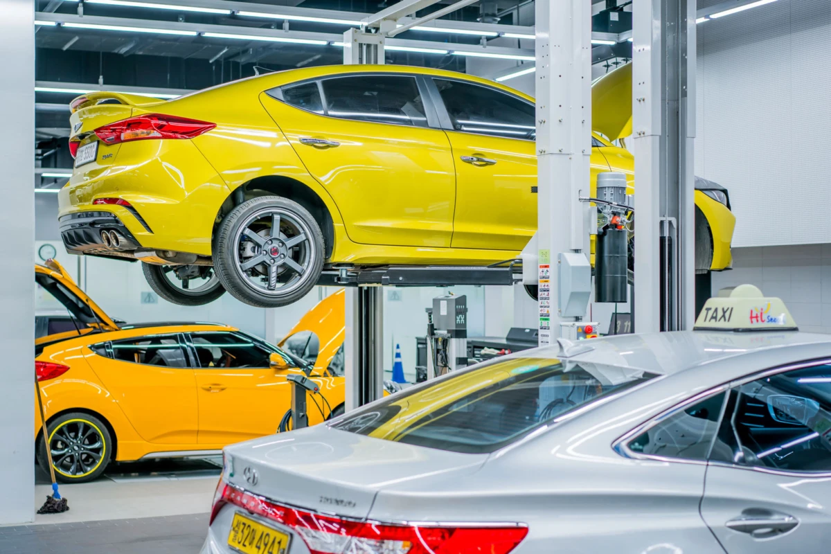 Yellow sedan elevated on a two-post car lift in an auto repair shop; another yellow sedan and a silver taxi visible nearby.