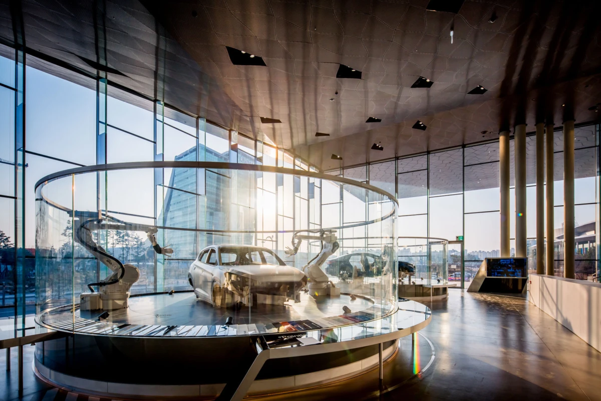 White sedan inside a circular glass enclosure surrounded by robotic arms in a modern car showroom.