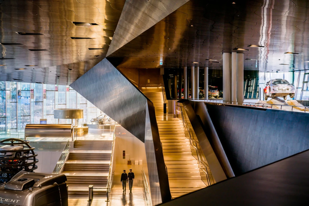 Two individuals walking in a modern car showroom atrium with escalators, steel architecture, and a car on a display platform.