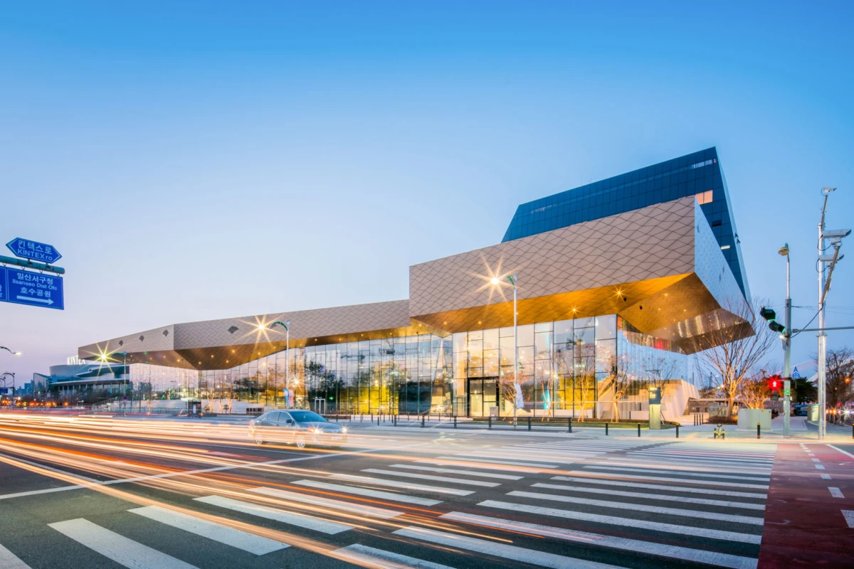 Geometric glass building at a busy intersection; long-exposure car lights streak across the zebra crosswalk.
