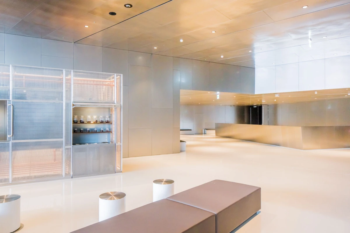 Modern stainless-steel lobby interior with curved counter, glass-front cabinet of jars, and white seating; empty.