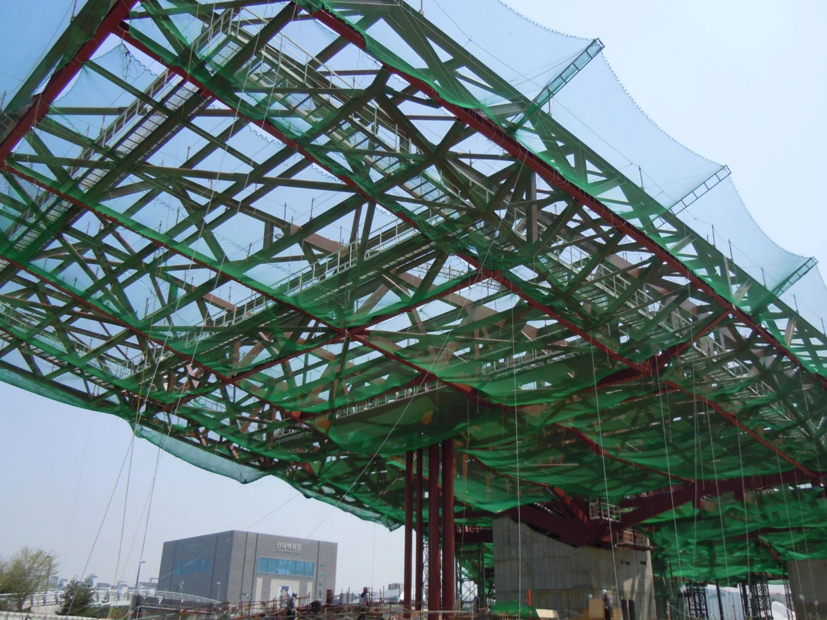 Green safety nets drape a red-columned steel framework under construction at an outdoor site.