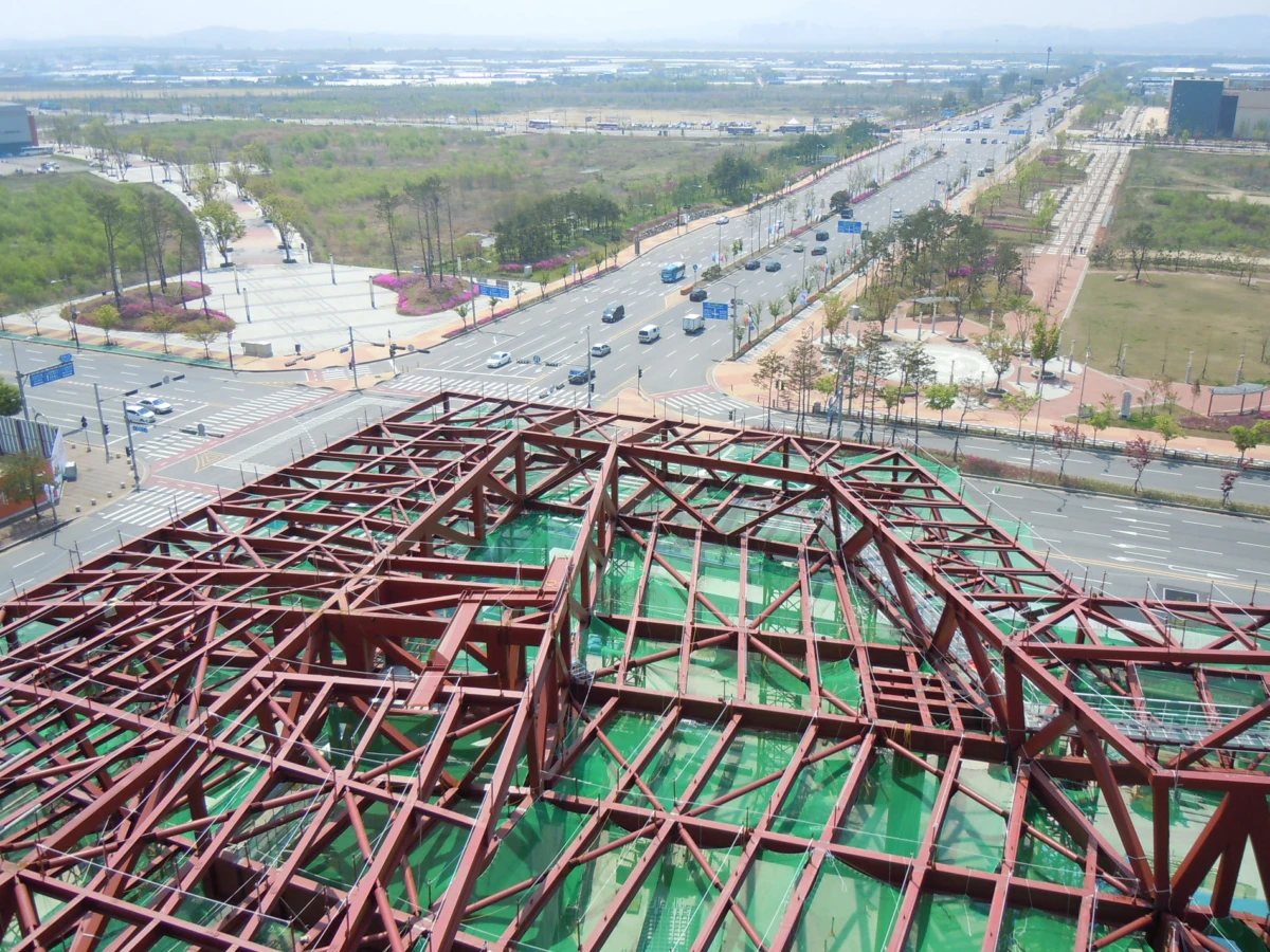 Aerial view of a red steel lattice roof under construction above a green surface, overlooking a busy multi-lane intersection with trees and parkland.
