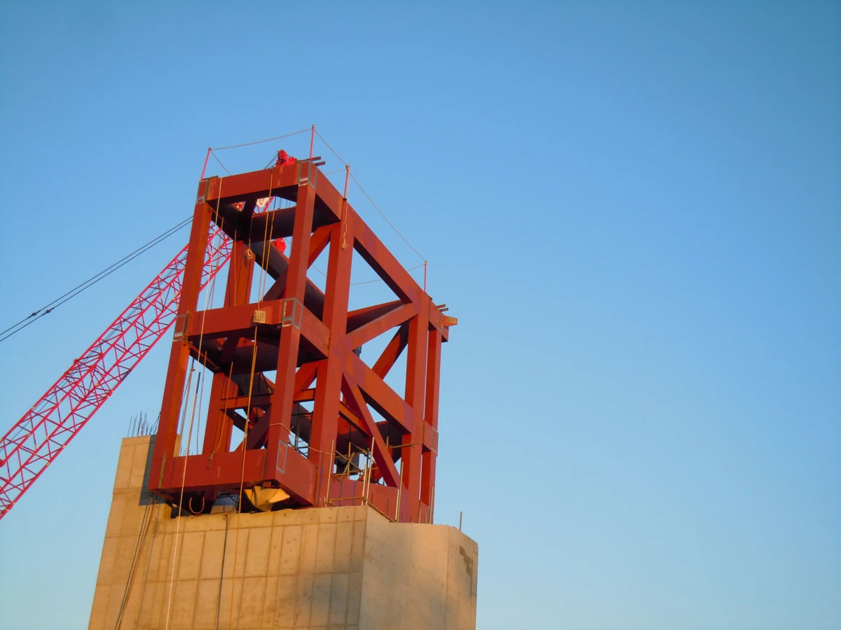 Red steel lattice tower rising from a concrete foundation; crane to the left, worker on top against a clear blue sky.