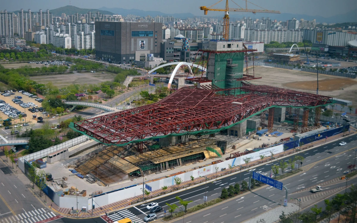 Large steel-framed canopy under construction with red lattice beams and a crane in an urban cityscape.
