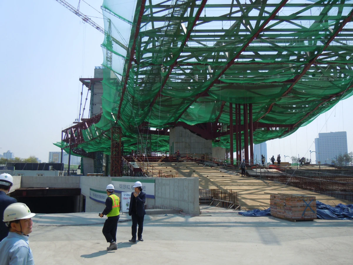 Construction site under green netting over a steel framework; workers in hard hats walk on concrete with stacked bricks nearby.