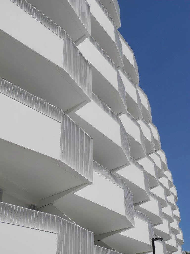 White high-rise residential building with staggered balconies and vertical railings against a clear blue sky.