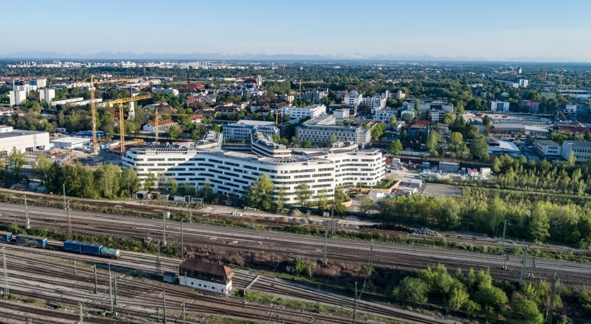 Aerial view of a curved white building under construction, with cranes, beside railway tracks in a suburban city.