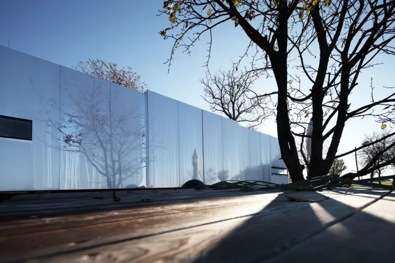 Reflective metal wall panels along a building mirror leafless trees; wooden deck foreground and clear blue sky.