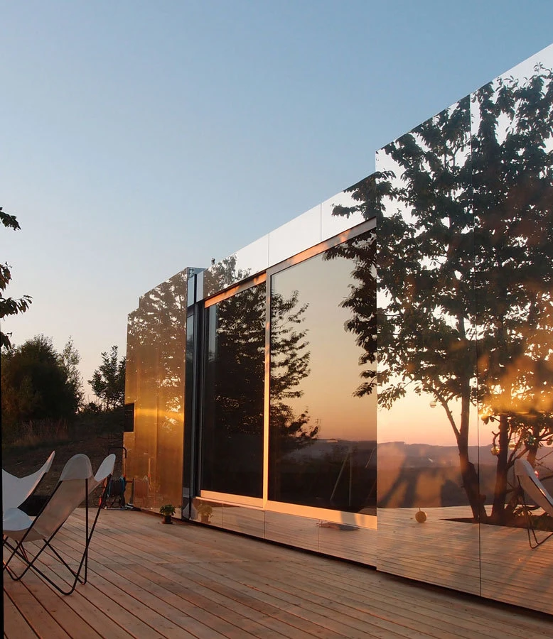 Modern glass-walled building on a wooden deck, reflecting sunset and trees; white outdoor lounge chairs nearby.