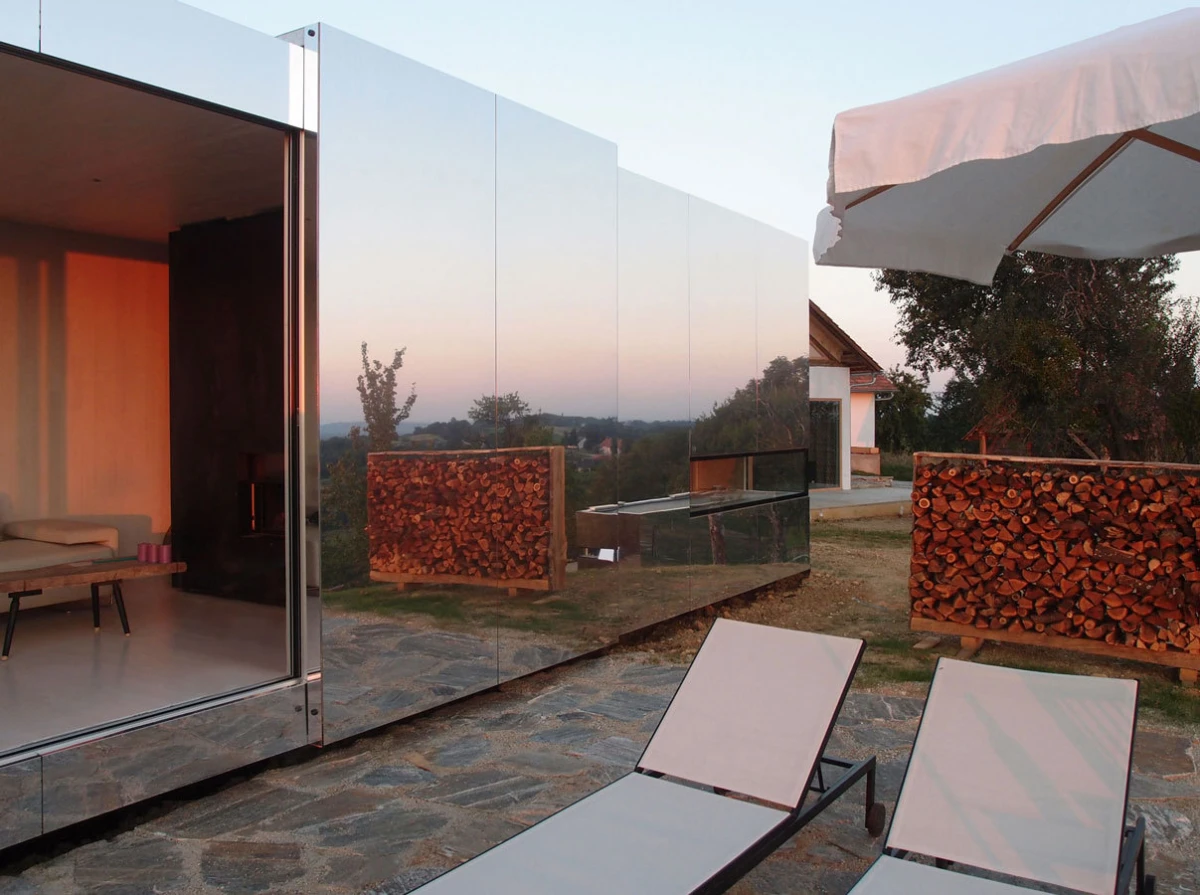 Modern glass-walled house reflecting the landscape; outdoor patio with two sun loungers, stacked firewood, and a large umbrella at dusk.