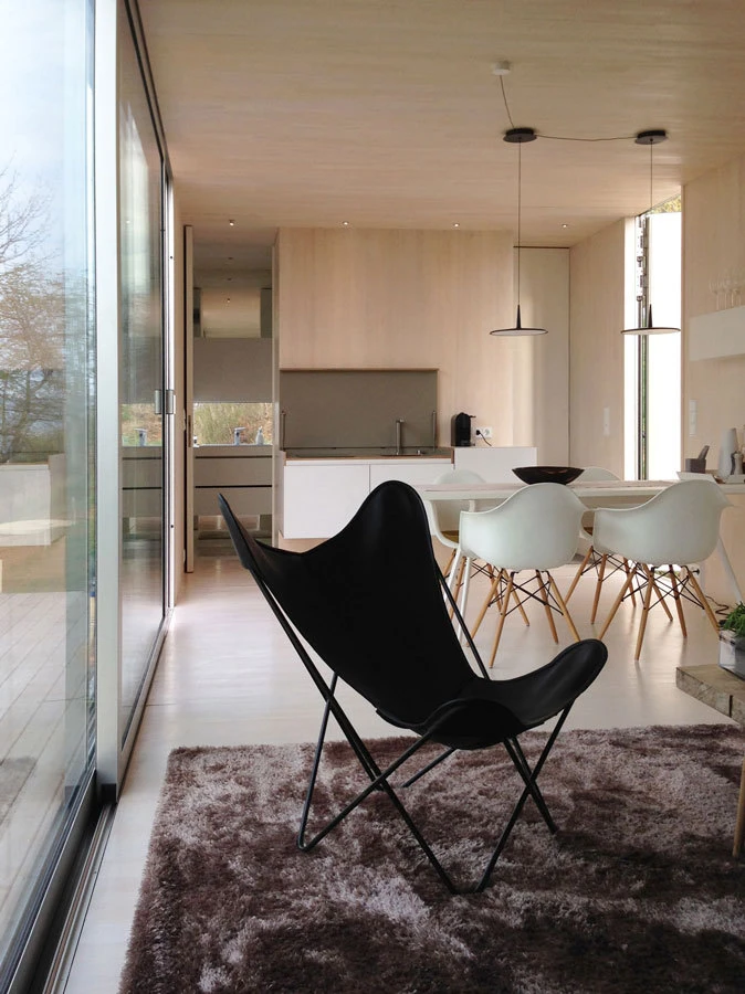 Black butterfly chair resting on a shaggy rug in an open-plan kitchen and dining area with glass walls and light wood furnishings.