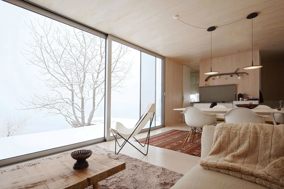 Bright minimalist living room with floor-to-ceiling windows overlooking a snowy landscape; butterfly chair, beige sofa with fur throw, white dining chairs around a table.