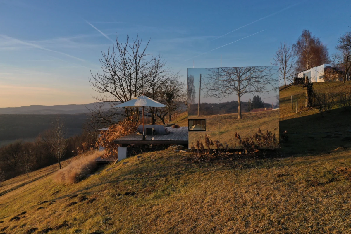 Mirrored cube sculpture on hillside deck with umbrella, reflecting surrounding trees at sunset.