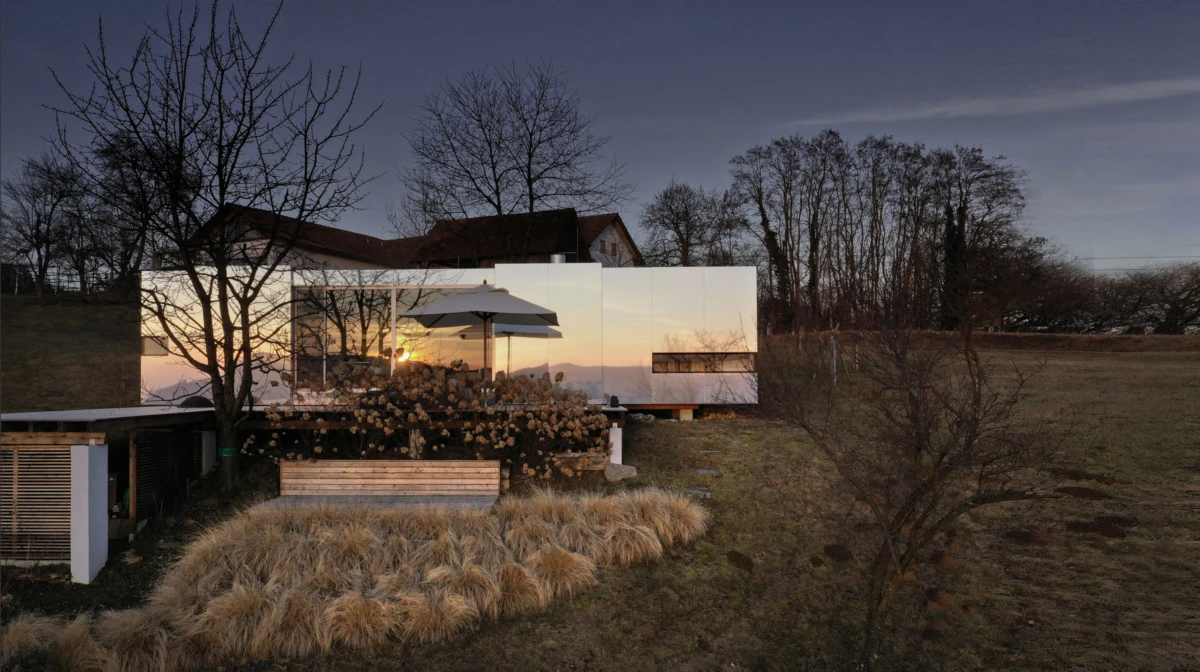 Contemporary glass-walled house reflecting sunset, with outdoor seating under a patio umbrella in a grassy rural yard with bare trees.