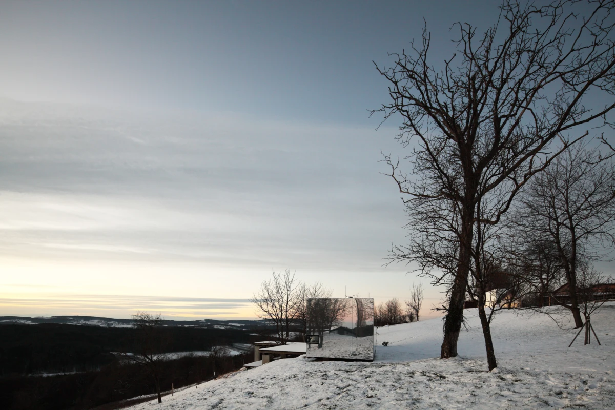 Reflective rectangular sculpture on a snow-covered hillside, reflecting the snow and bare trees at sunset.
